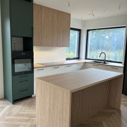 BIASTYL meble na wymiar - Nowoczesna kuchnia w trakcie montażu z zielonymi szafkami, drewnianymi frontami i wyspą z lamelami, widoczne wiszące kable od oświetlenia.
