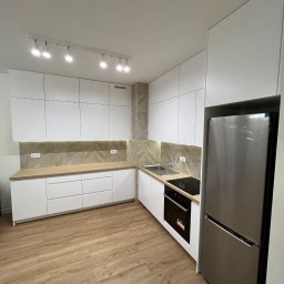 BIASTYL meble na wymiar - Nowoczesna kuchnia z białymi, gładkimi frontami szafek, drewnianym blatem i marmurkową ścianą nad blatem, oświetlona listwą z reflektorami.