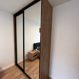 BIASTYL meble na wymiar - Szafa wnękowa z lustrzanymi drzwiami przesuwnymi, obudowa z drewna w ciepłym odcieniu, czarne ramy drzwi, widoczna szafka nocna i fragment łóżka w odbiciu.