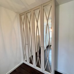 BIASTYL meble na wymiar - Czterodrzwiowa szafa wnękowa z lustrami i dekoracyjnymi wycięciami w kształcie falistych linii, zamontowana we wnęce pomiędzy białymi ścianami i ciemną podłogą.