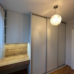 BIASTYL meble na wymiar - Szara szafa przesuwna z drewnianym blatem biurka wbudowanym w jej część, oświetlona dekoracyjna lampa sufitowa.