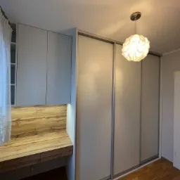 Szara szafa przesuwna z drewnianym blatem biurka wbudowanym w jej część, oświetlona dekoracyjna lampa sufitowa.