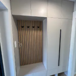 Nowoczesna szafa wnękowa w przedpokoju z drewnianymi lamelami na ścianie wnęki i czarnymi haczykami na ubrania.