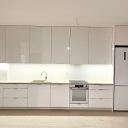 Minimalistyczna, biała kuchnia z drewnianym blatem, złotą baterią i wbudowanym sprzętem AGD, oświetlona listwą LED pod szafkami wiszącymi.