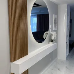 Nowoczesny przedpokój z owalnym lustrem podświetlanym LED, drewnianymi lamelami, białą konsolą z szufladami i marmurową podłogą.