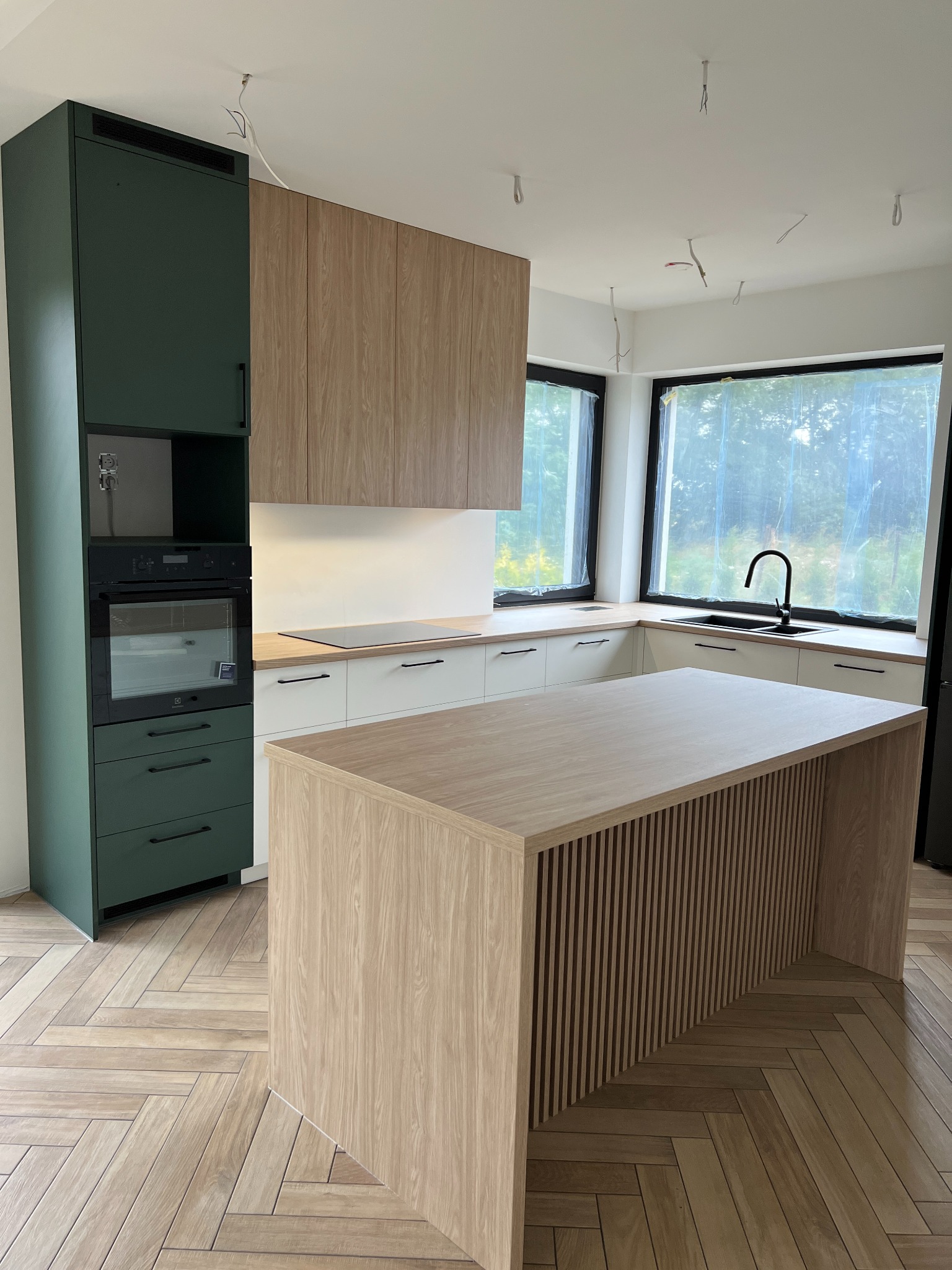 Nowoczesna kuchnia w trakcie montażu z zielonymi szafkami, drewnianymi frontami i wyspą z lamelami, widoczne wiszące kable od oświetlenia.