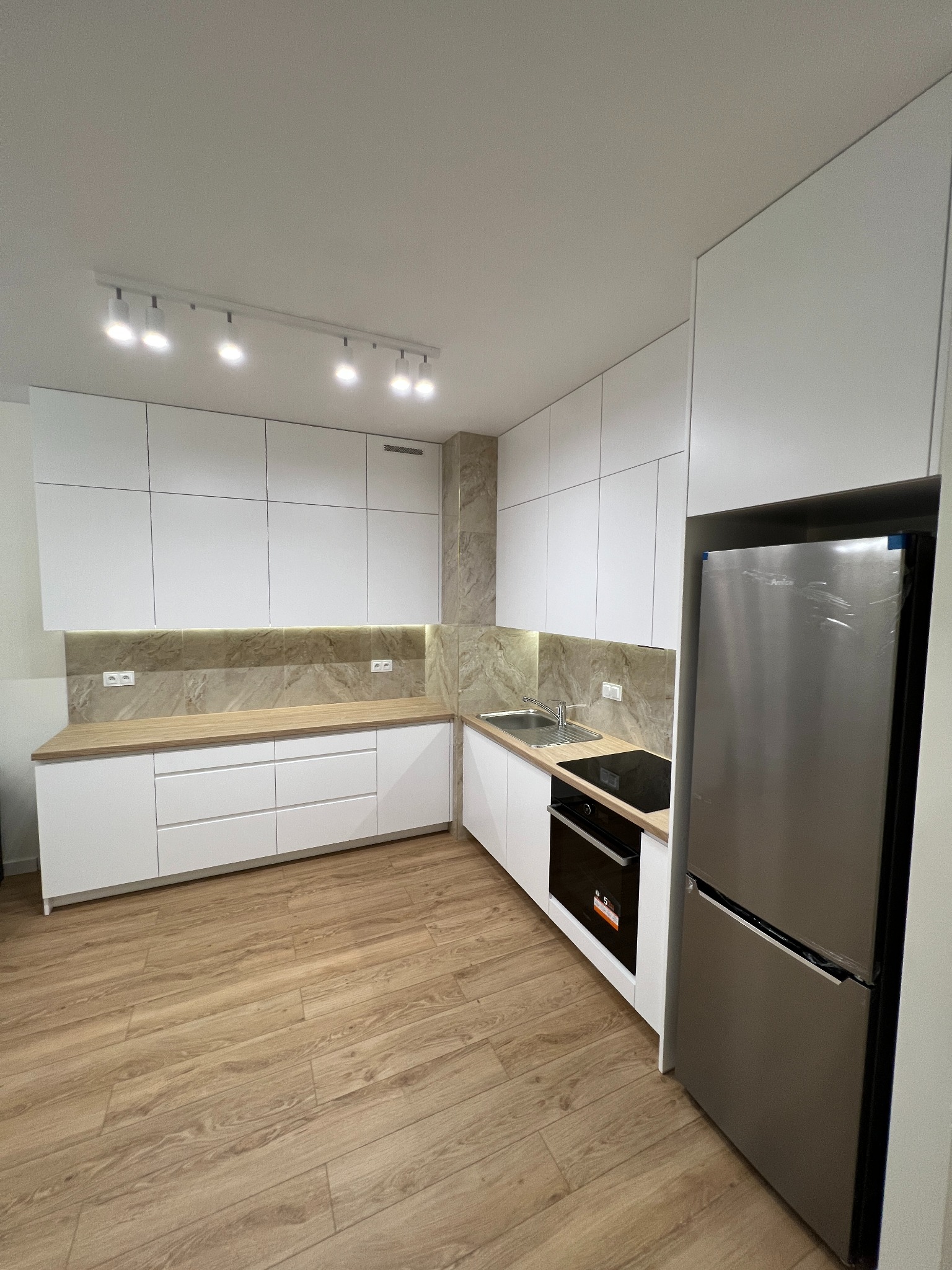 Nowoczesna kuchnia z białymi, gładkimi frontami szafek, drewnianym blatem i marmurkową ścianą nad blatem, oświetlona listwą z reflektorami.