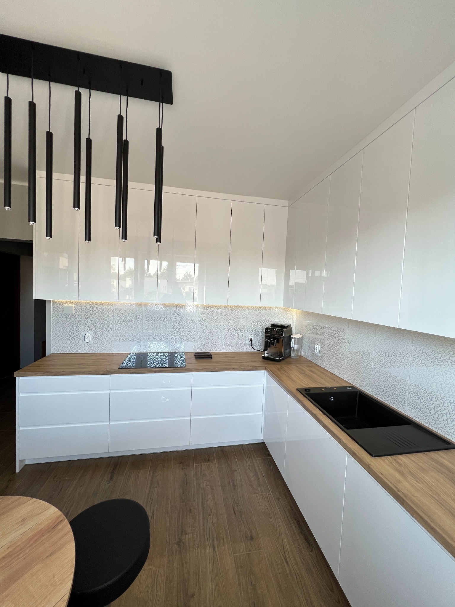 Nowoczesna kuchnia z białymi, lakierowanymi frontami szafek, drewnianym blatem i czarnymi, wiszącymi lampami tubami. Na blacie ekspres do kawy. Podświetlenie LED nad blatem.