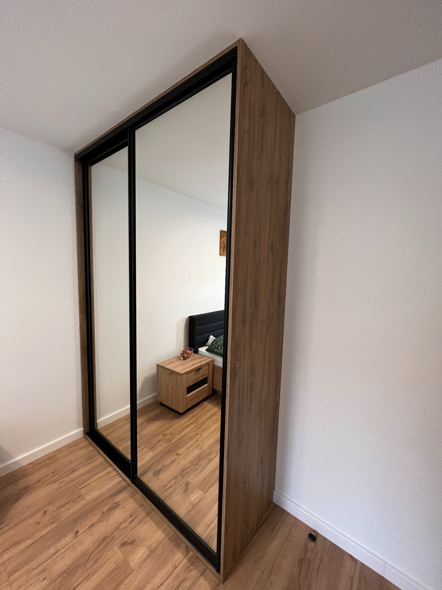 Szafa wnękowa z lustrzanymi drzwiami przesuwnymi, obudowa z drewna w ciepłym odcieniu, czarne ramy drzwi, widoczna szafka nocna i fragment łóżka w odbiciu.