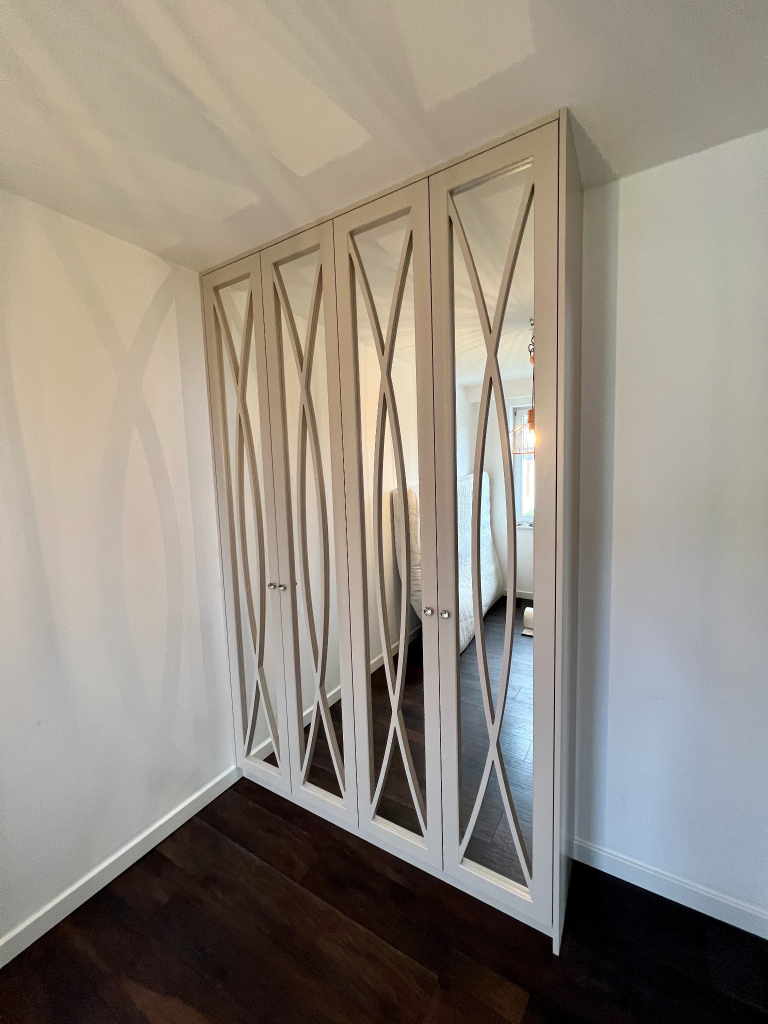 Czterodrzwiowa szafa wnękowa z lustrami i dekoracyjnymi wycięciami w kształcie falistych linii, zamontowana we wnęce pomiędzy białymi ścianami i ciemną podłogą.