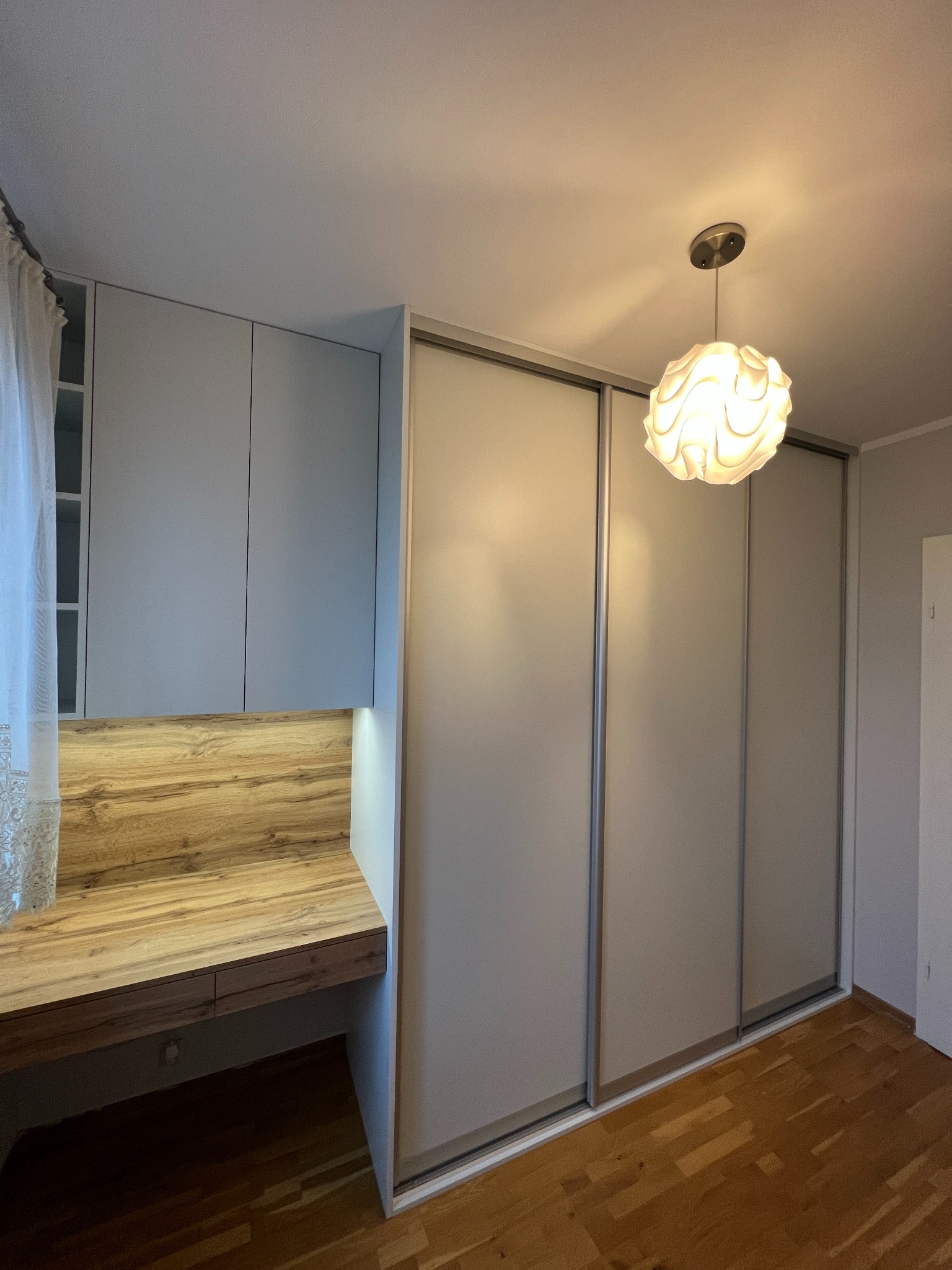 Szara szafa przesuwna z drewnianym blatem biurka wbudowanym w jej część, oświetlona dekoracyjna lampa sufitowa.