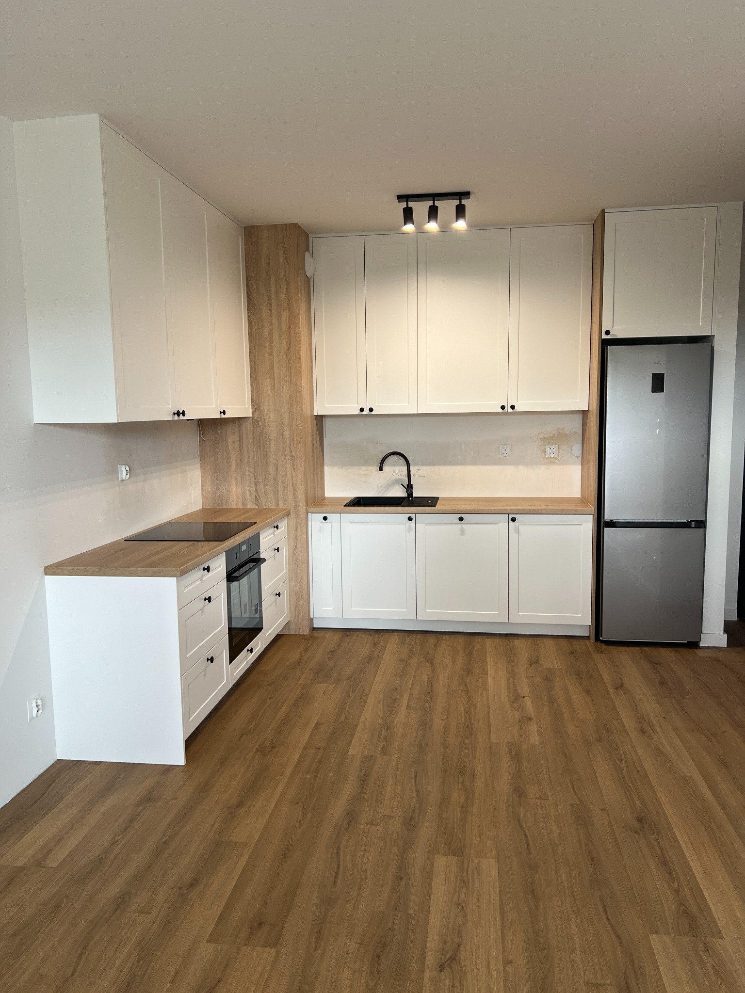 Nowoczesna kuchnia z białymi szafkami, drewnianymi blatami i czarnymi akcentami, w tym bateria i oświetlenie sufitowe. Widoczne niedokończone ściany nad blatem.