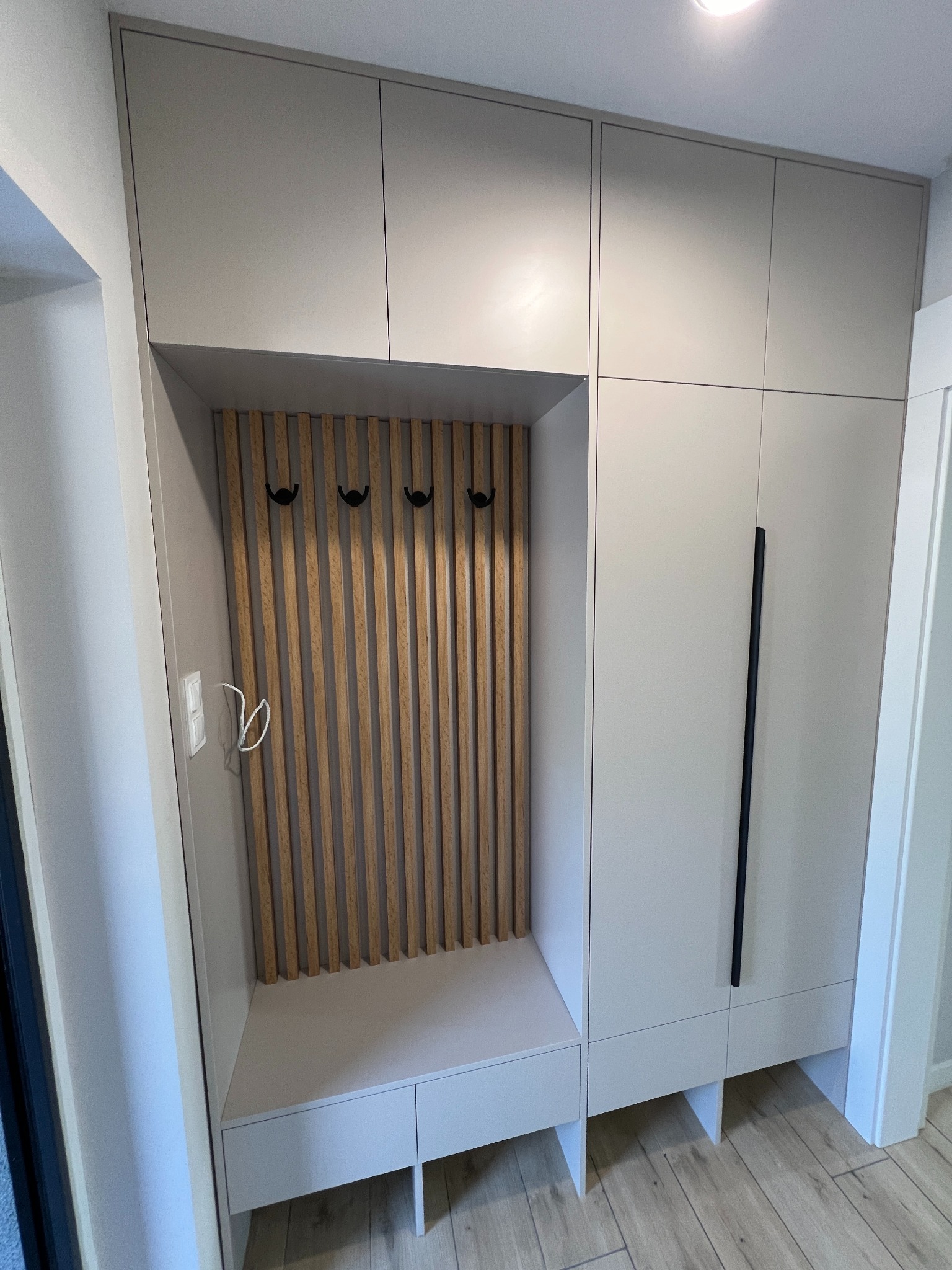 Nowoczesna szafa wnękowa w przedpokoju z drewnianymi lamelami na ścianie wnęki i czarnymi haczykami na ubrania.