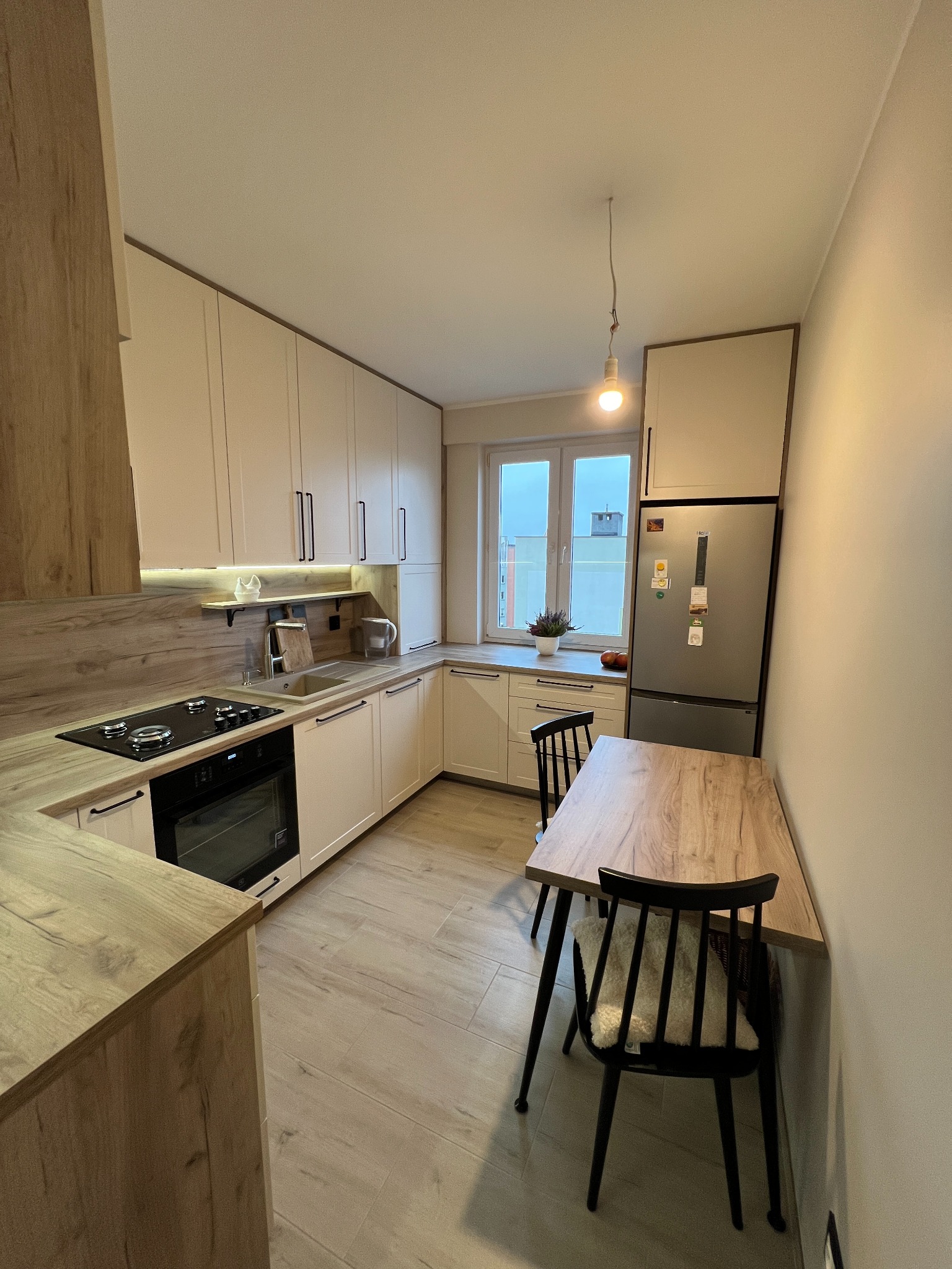 Jasna kuchnia w Baciutach z drewnianymi blatami i czarnymi uchwytami, uzupełniona o stół z krzesłami w podobnym stylu, oświetlona naturalnym światłem z okna.