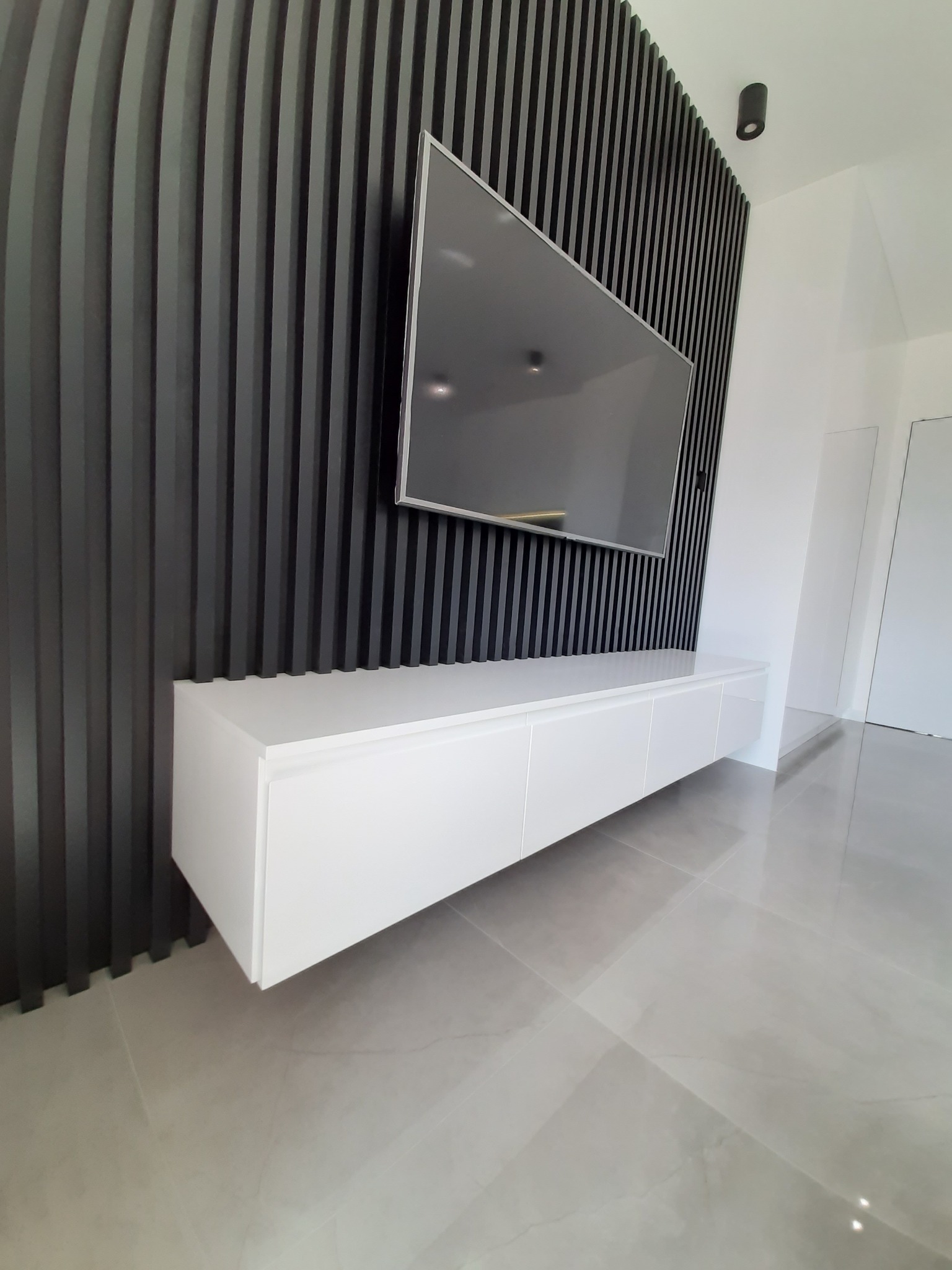 Nowoczesna szafka RTV w kolorze białym, zawieszona pod telewizorem na czarnej ścianie z pionowych paneli dekoracyjnych, na podłodze jasne płytki. Minimalistyczny design wnętrza.