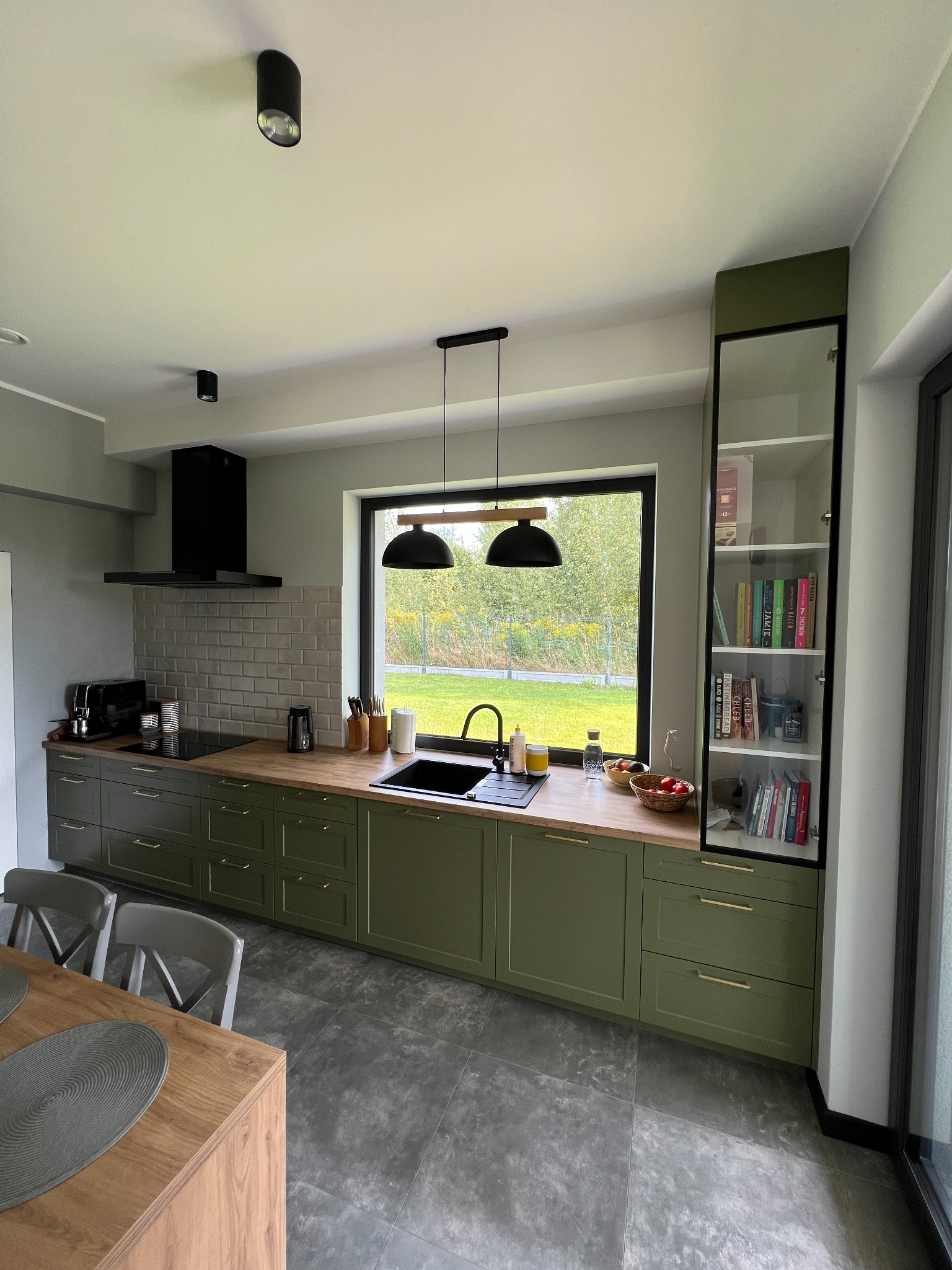 Nowoczesna kuchnia z oliwkowozielonymi frontami szafek, drewnianym blatem i dużym oknem z widokiem na zieleń. Nad blatem wiszą dwie czarne lampy. Po prawej stronie wysoka witryna z książkami.