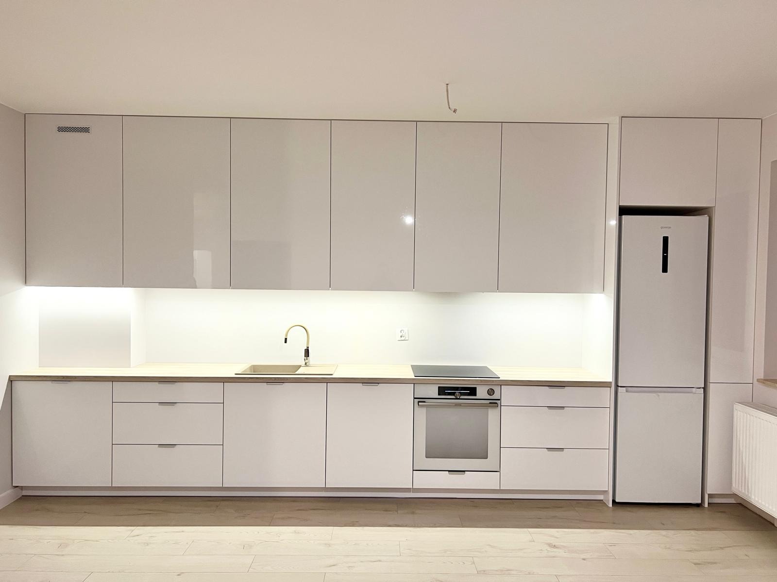 Minimalistyczna, biała kuchnia z drewnianym blatem, złotą baterią i wbudowanym sprzętem AGD, oświetlona listwą LED pod szafkami wiszącymi.