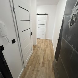 BDR-BUD Sp. Z O.O - Korytarz po remoncie z drewnopodobną podłogą, białymi drzwiami z czarnymi akcentami i szarą, zabezpieczoną folią, wejściową. Nowoczesny design.