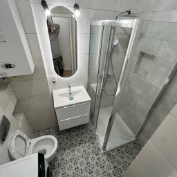 BDR-BUD Sp. Z O.O - Wykończona łazienka z kabiną prysznicową, umywalką z szafką i toaletą. Na podłodze wzorzyste płytki. W lustrze odbija się osoba robiąca zdjęcie.