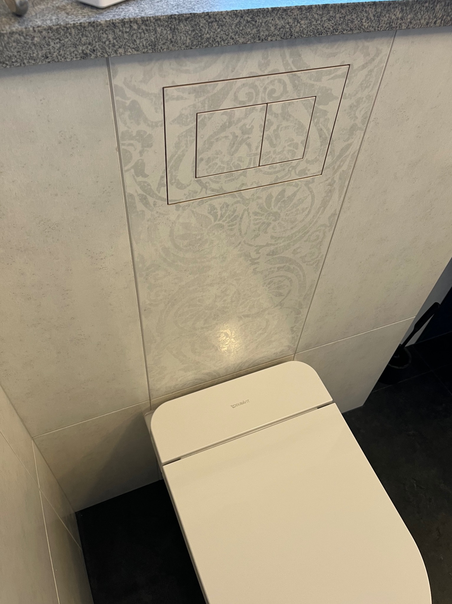 Nowoczesna toaleta Duravit z ukrytym spłukiwaniem w ścianie wykończonej płytkami z delikatnym, kwiatowym wzorem i szarą listwą przypodłogową.