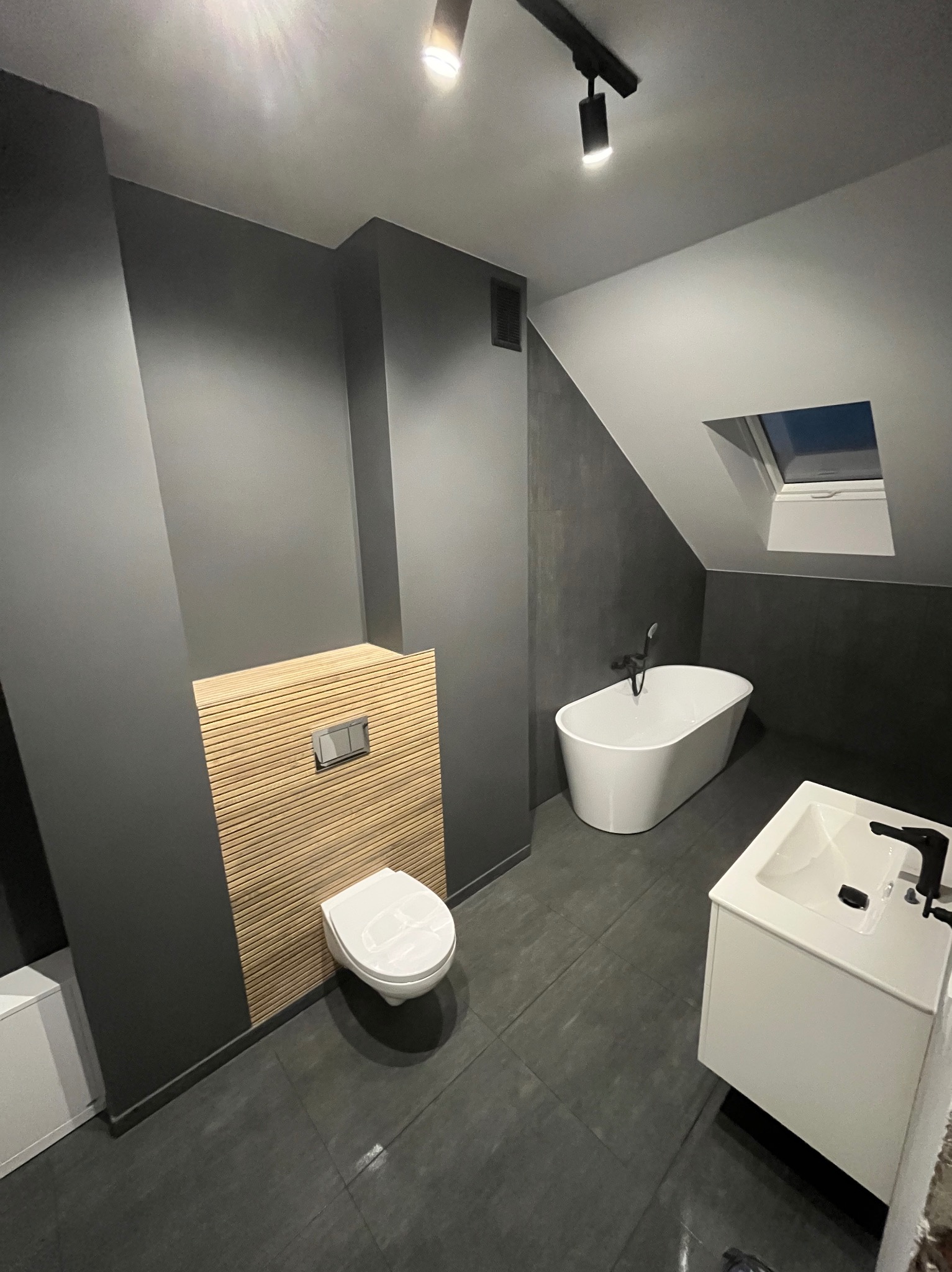 Nowoczesna łazienka na poddaszu z szarą kolorystyką ścian, drewnianym panelem dekoracyjnym za toaletą, białą wanną wolnostojącą i umywalką z szafką.