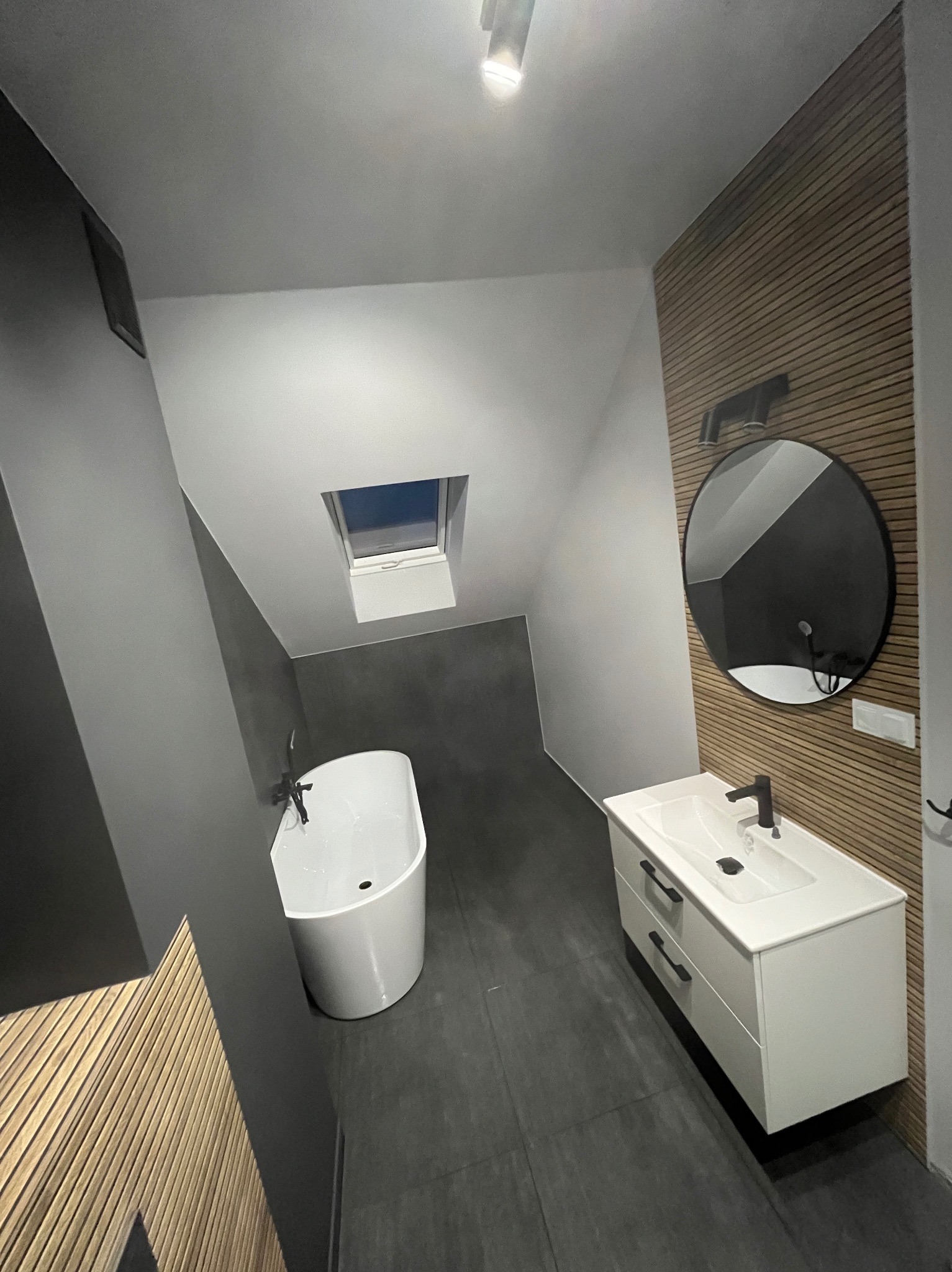 Nowoczesna łazienka na poddaszu z wolnostojącą wanną, umywalką z szafką i okrągłym lustrem na tle drewnianej okładziny ściennej, szare ściany i podłoga.