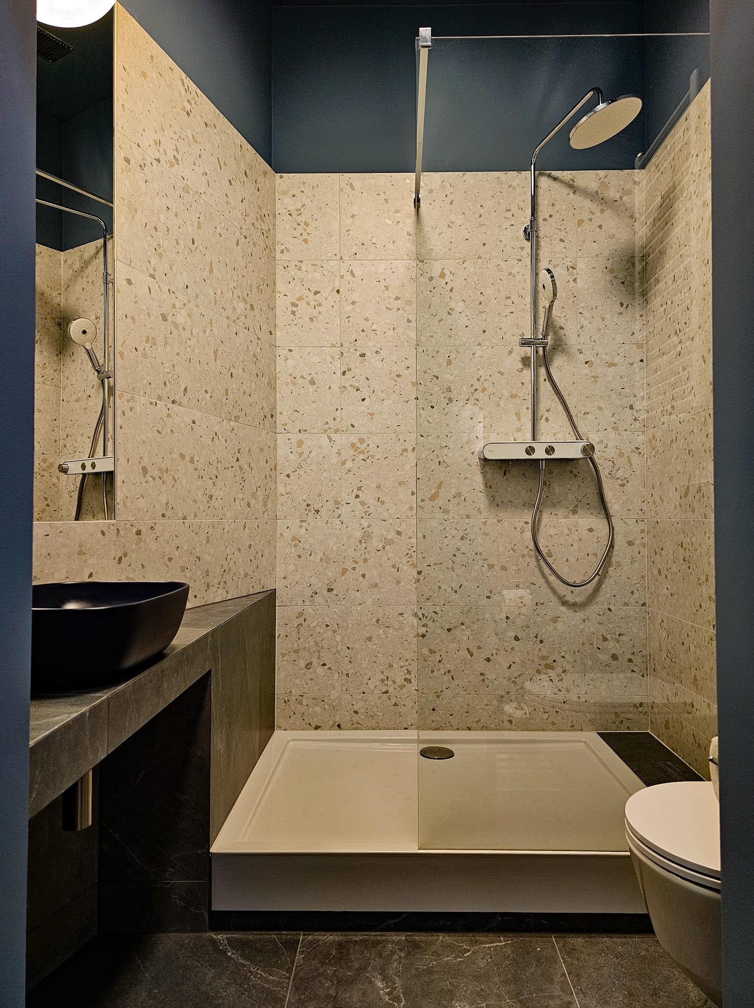 Nowoczesna łazienka z kabiną prysznicową typu walk-in, umywalką nablatową i płytkami imitującymi lastryko na ścianach, wykończona w ciemnej kolorystyce.