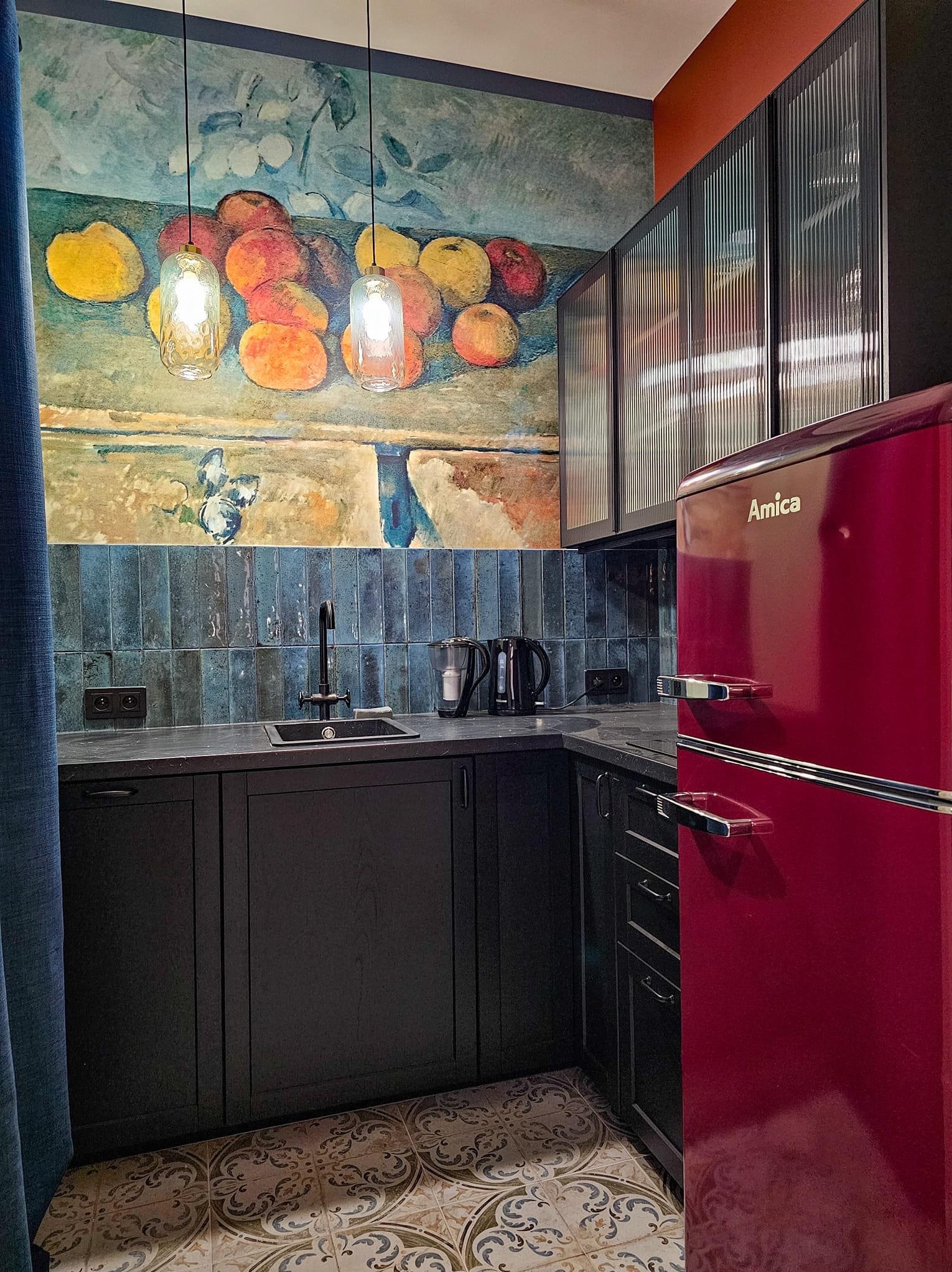 Kuchenne wnętrze z ciemnymi szafkami, mozaikową podłogą, błękitnymi płytkami nad blatem, obrazem owoców na ścianie i bordową lodówką Amica w stylu retro.