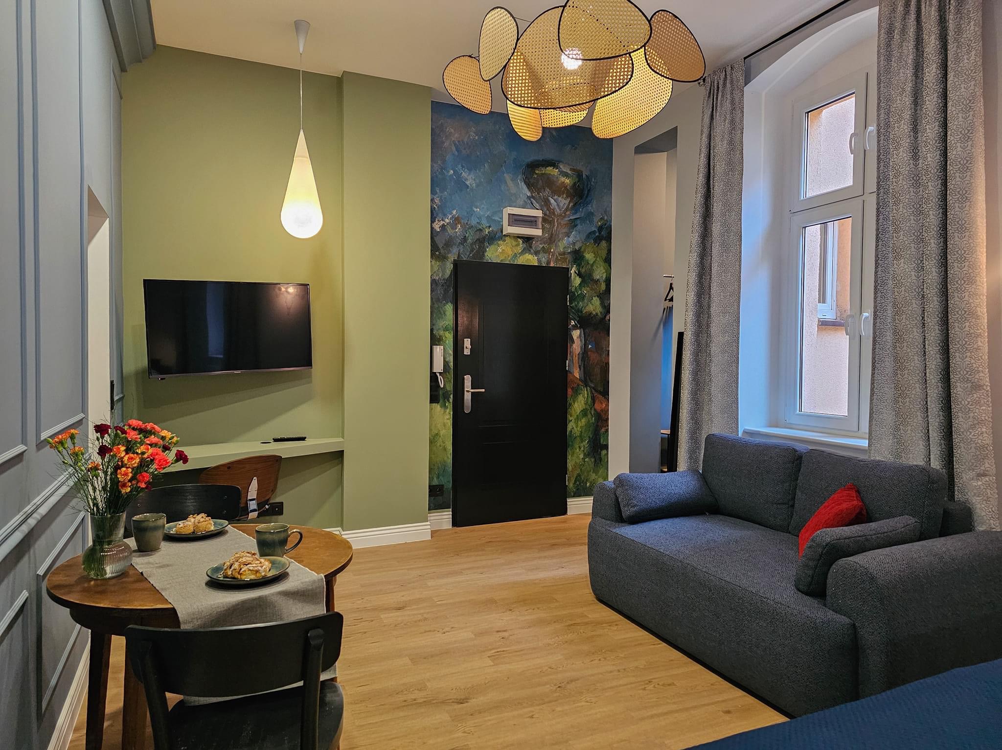 Jasne wnętrze mieszkania po remoncie: drewniana podłoga, zielone ściany, okrągły stół z kwiatami i ciastkami, szara sofa z czerwoną poduszką, oryginalna lampa sufitowa i malowidło na ścianie przy...