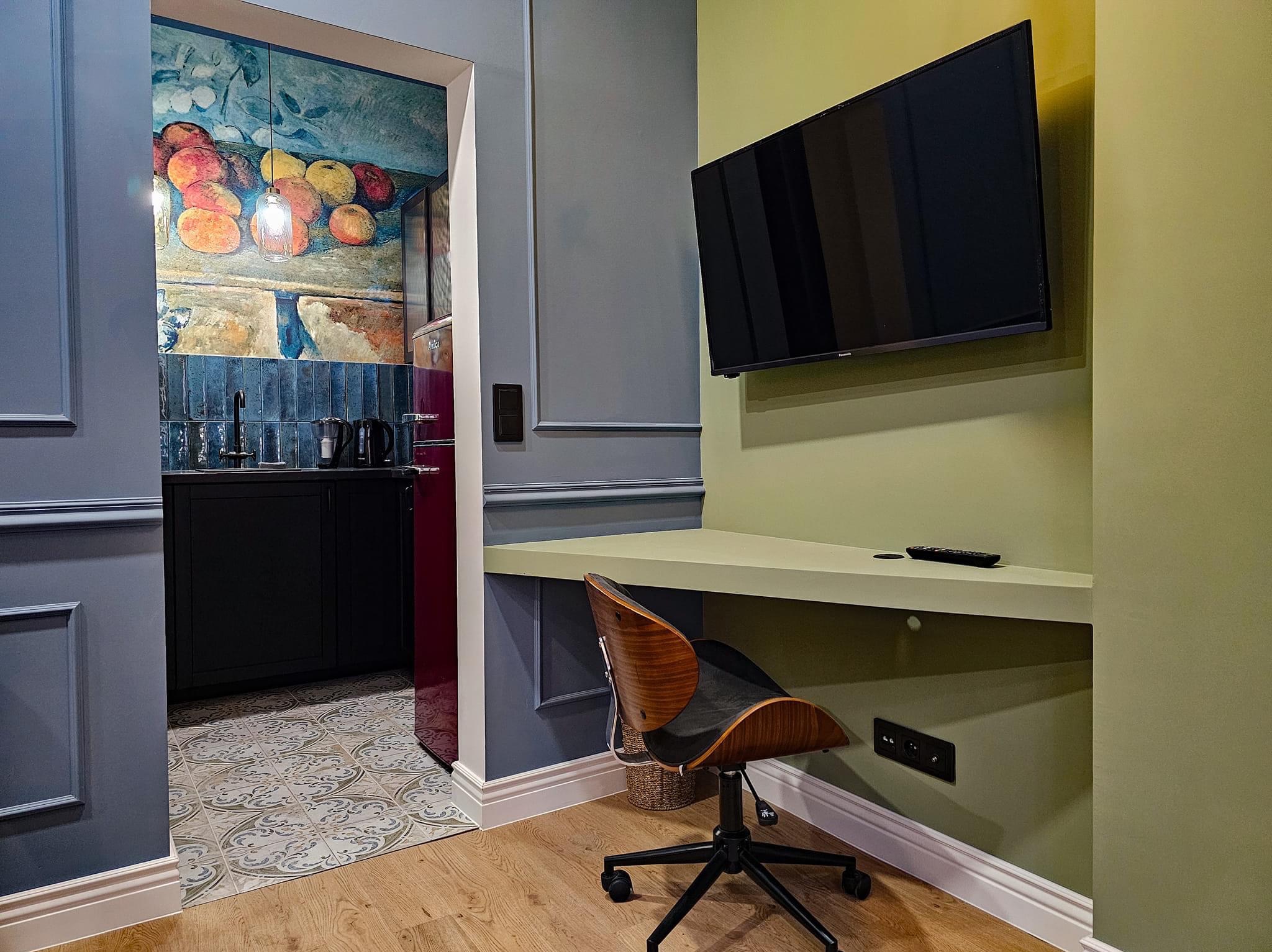 Nowoczesne wnętrze z biurkiem narożnym, telewizorem na ścianie w odcieniu oliwkowym, krzesłem biurowym z drewnianym oparciem i widokiem na kuchnię z motywem owoców na ścianie i turkusowymi kafelkami.