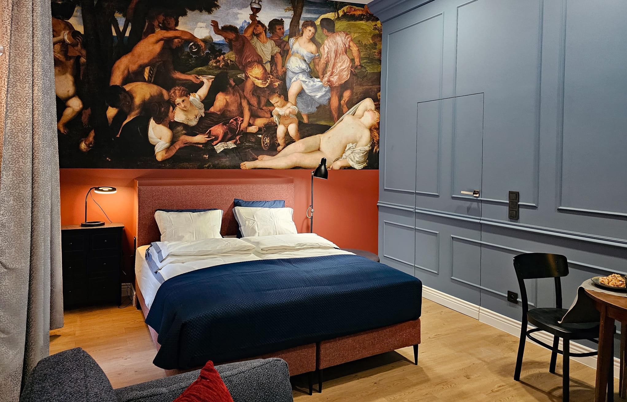 Sypialnia z łóżkiem małżeńskim, ciemnoniebieską narzutą i zagłówkiem w kolorze terakoty, nad którym widnieje malowidło ścienne w stylu renesansowym; ściany w kolorze gołębim z dekoracyjnymi listwami.