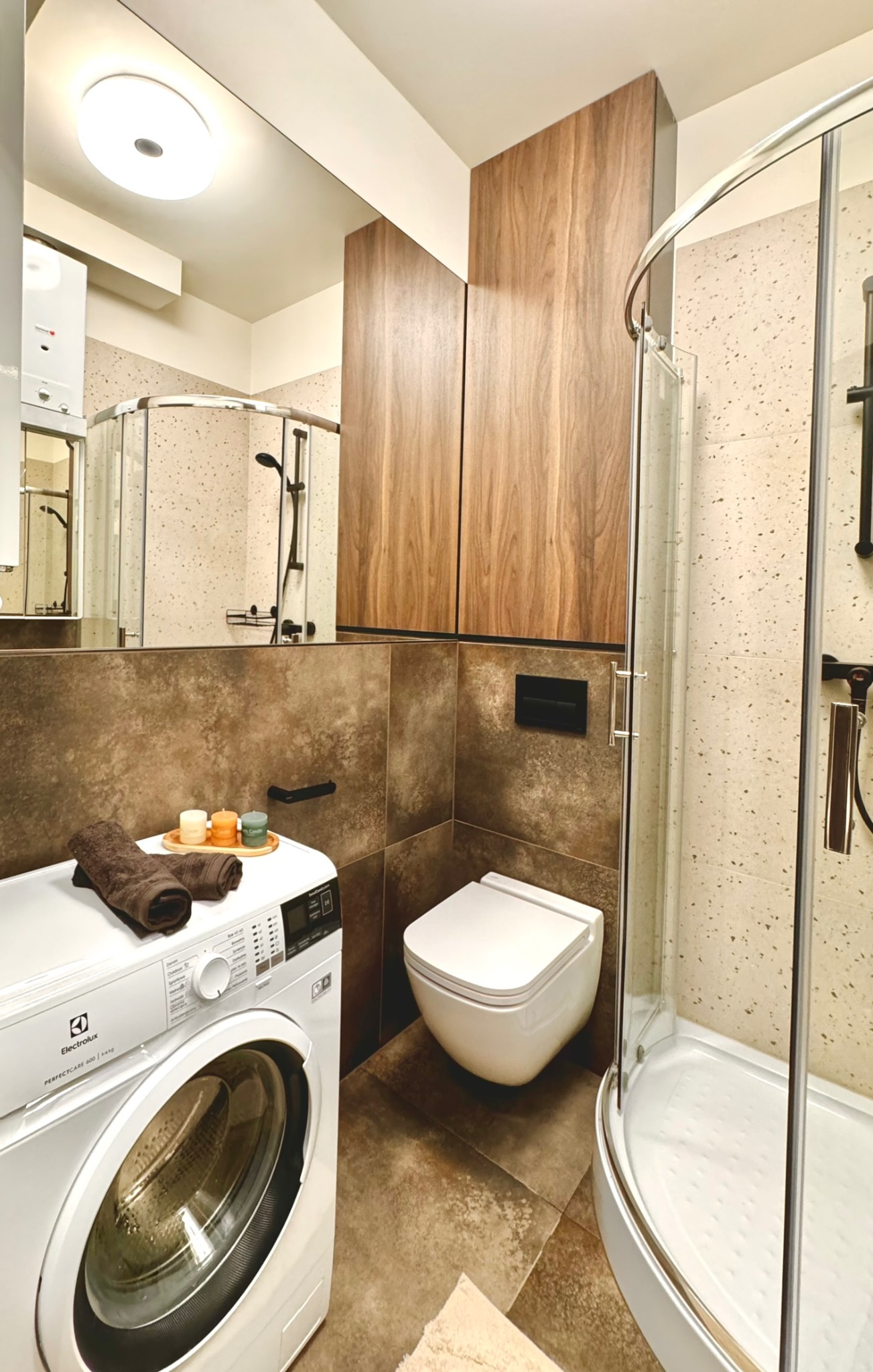 Nowoczesna łazienka z pralką Electrolux, wiszącą toaletą i kabiną prysznicową, wykończona brązowymi płytkami i drewnianymi szafkami.