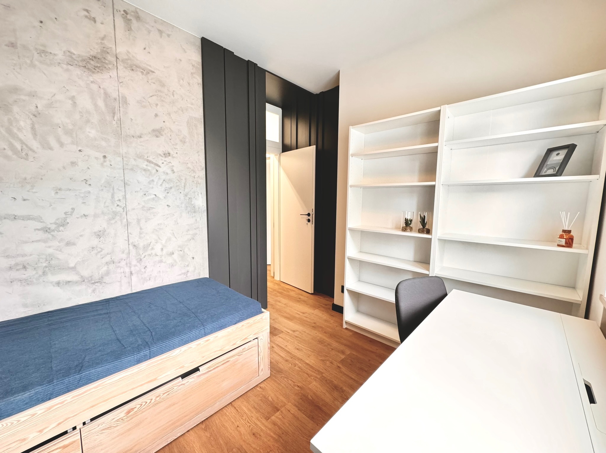 Nowoczesny pokój z łóżkiem z szufladami, ścianą o fakturze betonu, białym regałem i biurkiem, drewnianą podłogą oraz czarnymi panelami dekoracyjnymi.