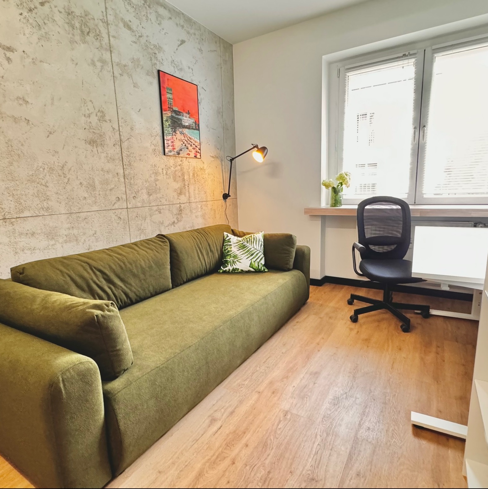 Jasne wnętrze pokoju z zieloną sofą, biurkiem i krzesłem na drewnianej podłodze, ściana z efektem betonu, oświetlona lampką biurkową.