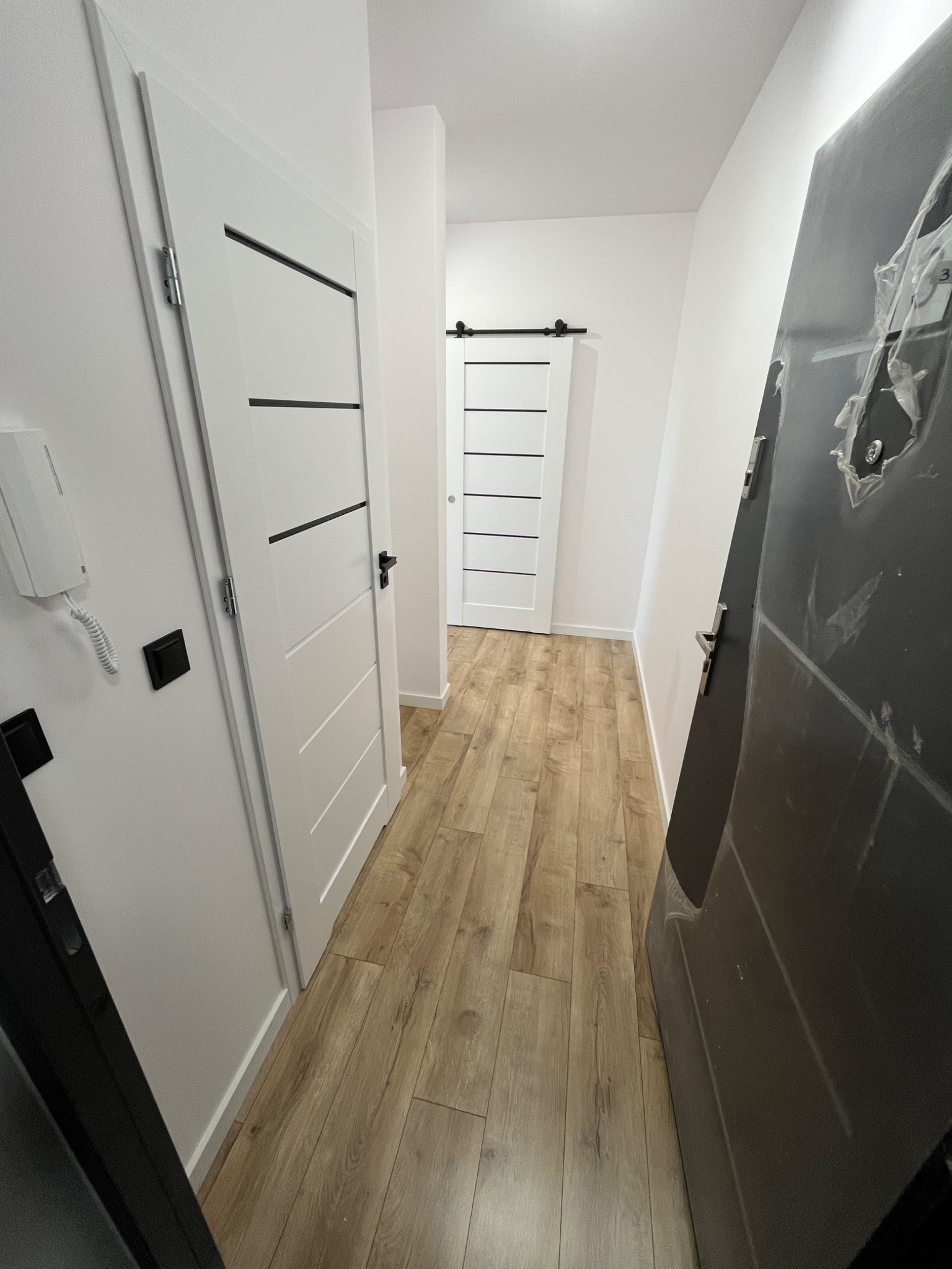 Korytarz po remoncie z drewnopodobną podłogą, białymi drzwiami z czarnymi akcentami i szarą, zabezpieczoną folią, wejściową. Nowoczesny design.