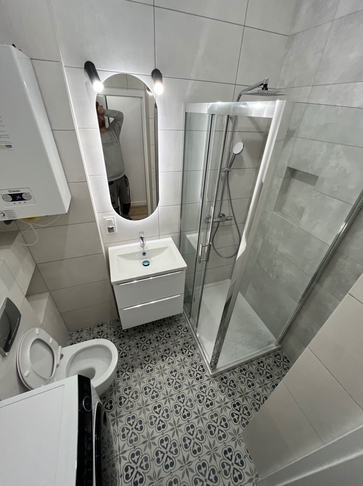 Wykończona łazienka z kabiną prysznicową, umywalką z szafką i toaletą. Na podłodze wzorzyste płytki. W lustrze odbija się osoba robiąca zdjęcie.