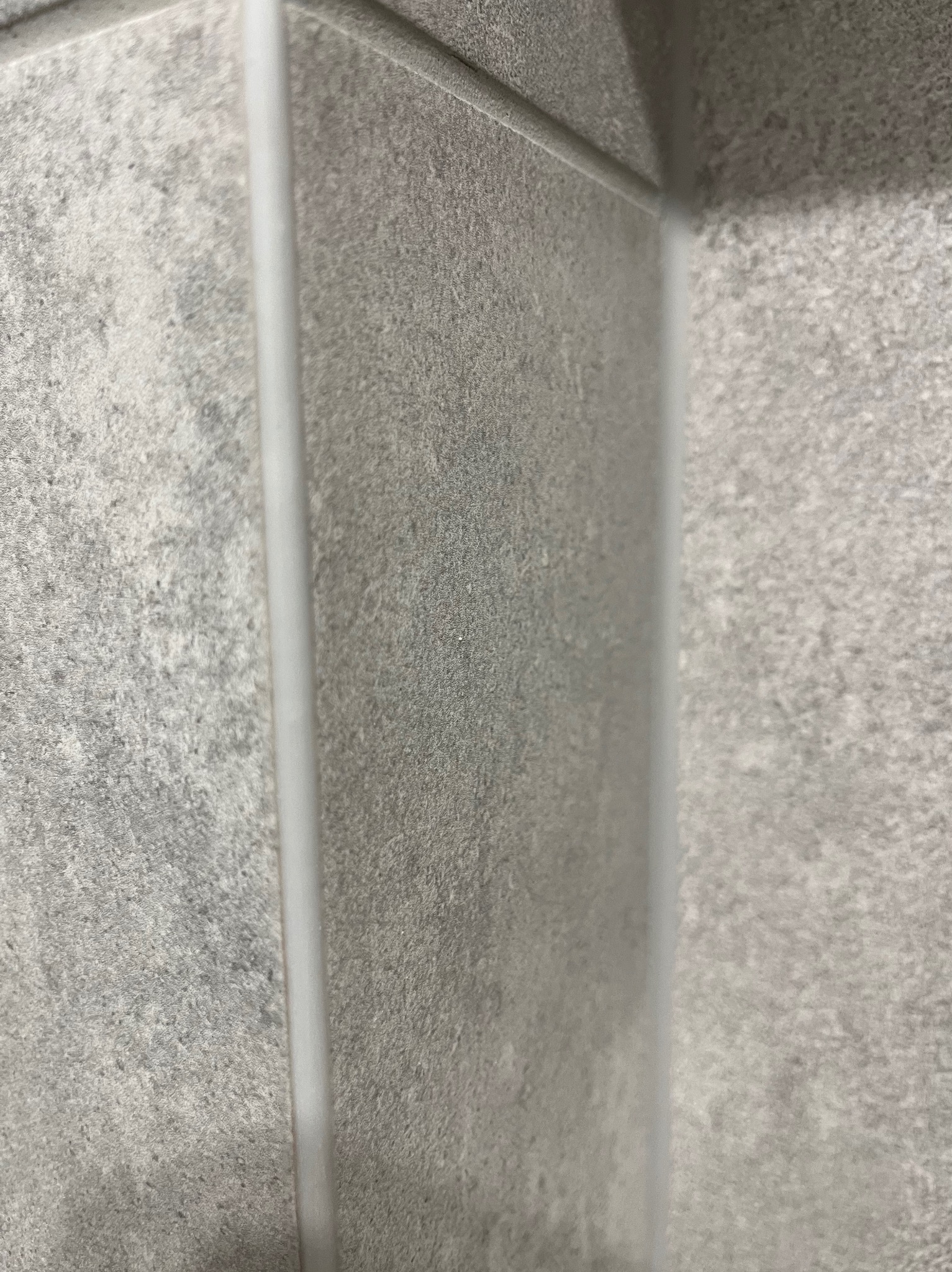 Szare, matowe płytki ceramiczne ułożone w rogu pomieszczenia, z widoczną białą fugą silikonową. Minimalistyczne wykończenie ściany, bliski kadr.