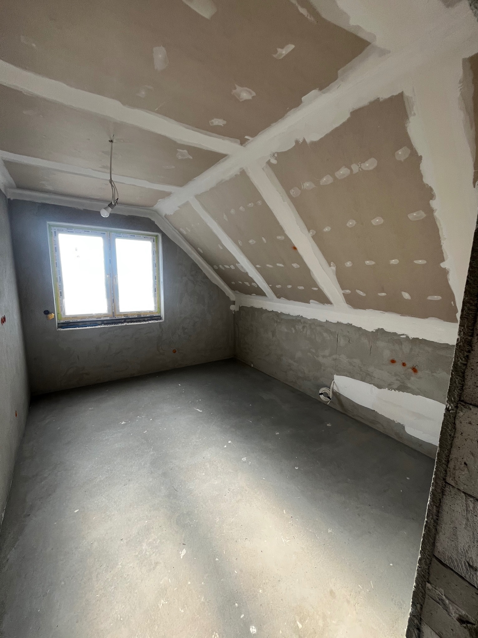Pokój na poddaszu w trakcie prac wykończeniowych: szpachlowane ściany, sufit z płyt karton-gips, okno. Widoczne niedoskonałości i surowy stan wnętrza.