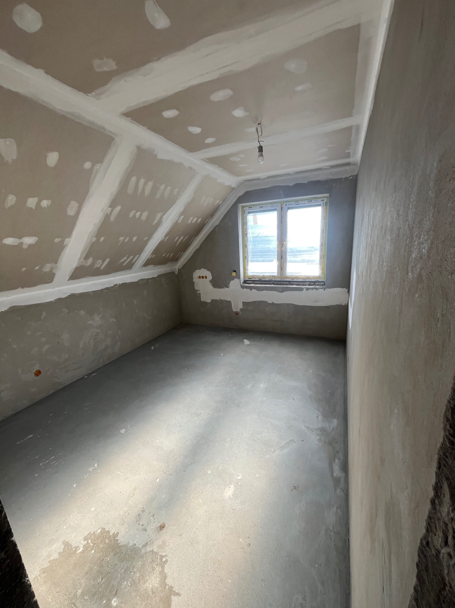Pokój na poddaszu w trakcie prac wykończeniowych: surowe ściany, płyty karton-gips na skosach sufitu, widoczne łączenia płyt, okno w tle, surowy beton na podłodze.