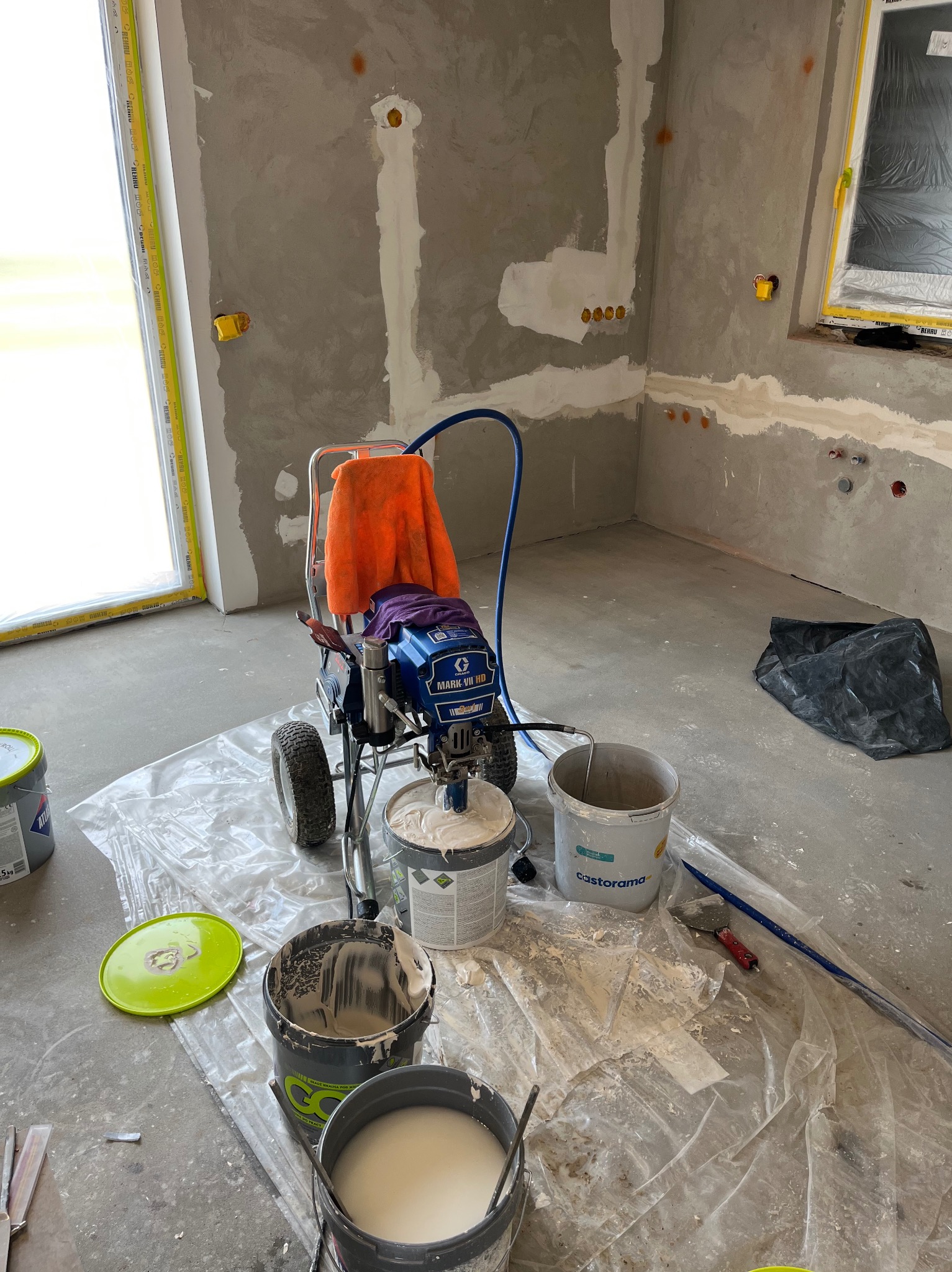 Przygotowania do malowania natryskowego w surowym wnętrzu: agregat malarski, wiadra z farbą, folia ochronna. Widoczne ślady szpachlowania na ścianach.