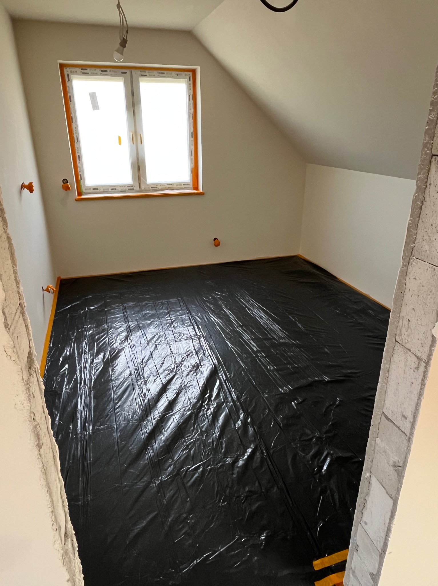 Pokój na poddaszu zabezpieczony czarną folią przed dalszymi pracami remontowymi. Widoczne listwy przypodłogowe i okno z pomarańczową ramą. Surowy stan wykończenia.