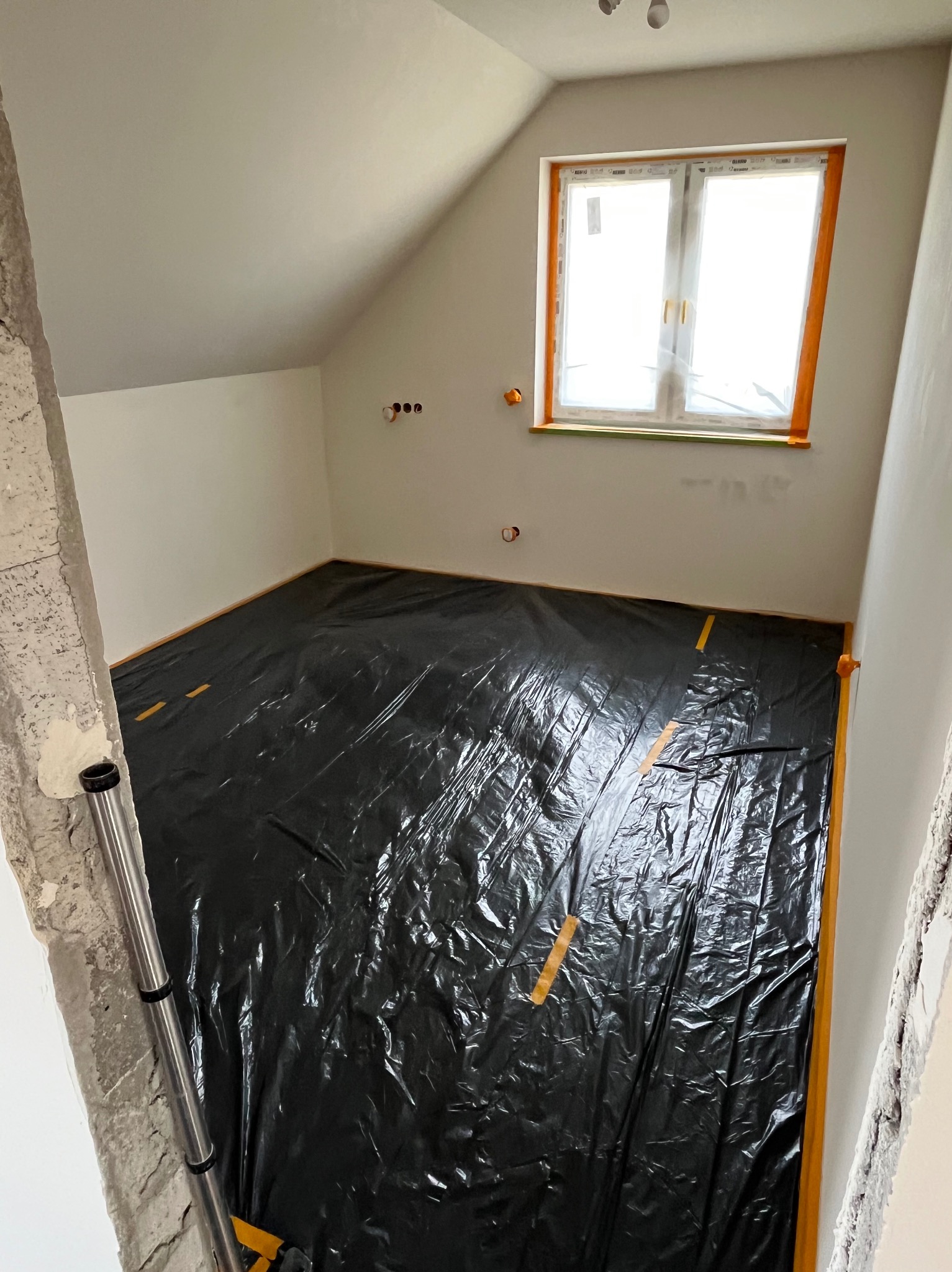 Pokój na poddaszu w trakcie remontu: zabezpieczona folią podłoga, okno z folią ochronną, ściany przygotowane do malowania. Widoczne gniazdka elektryczne i surowy fragment ściany.