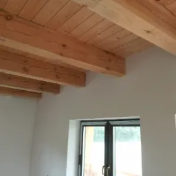 Drewniany sufit z belek w jasnym pomieszczeniu z dwoma oknami w czarnych ramach, widok z dołu.