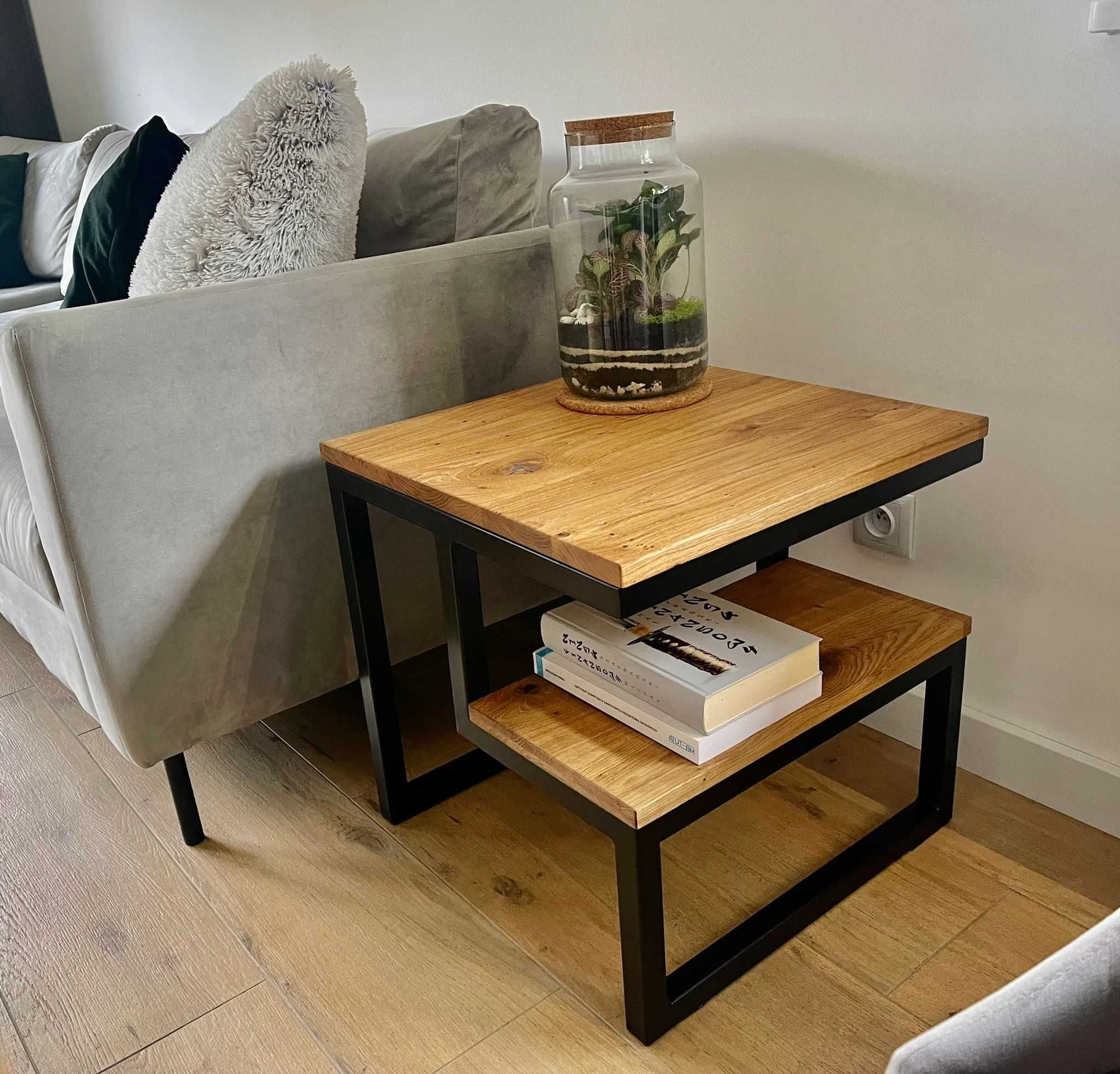 Nowoczesny stolik kawowy z drewnianym blatem i czarnymi metalowymi nogami, na którym stoi szklany słój z roślinami i leżą dwie książki, obok szarej sofy.