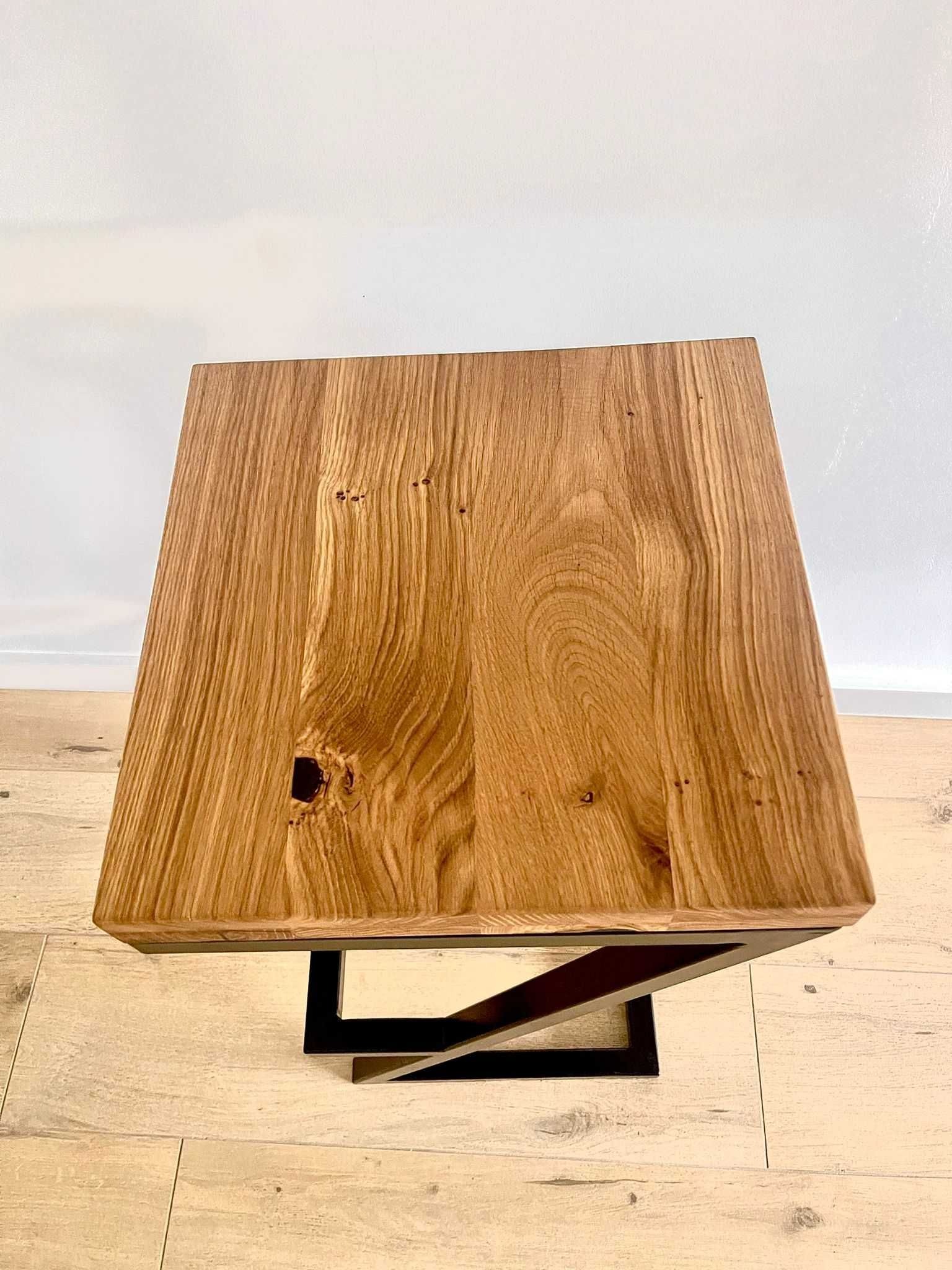 Stolik kawowy z blatem z litego drewna dębowego z widocznymi sękami i czarną, geometryczną podstawą z metalu, sfotografowany z góry na jasnej podłodze.