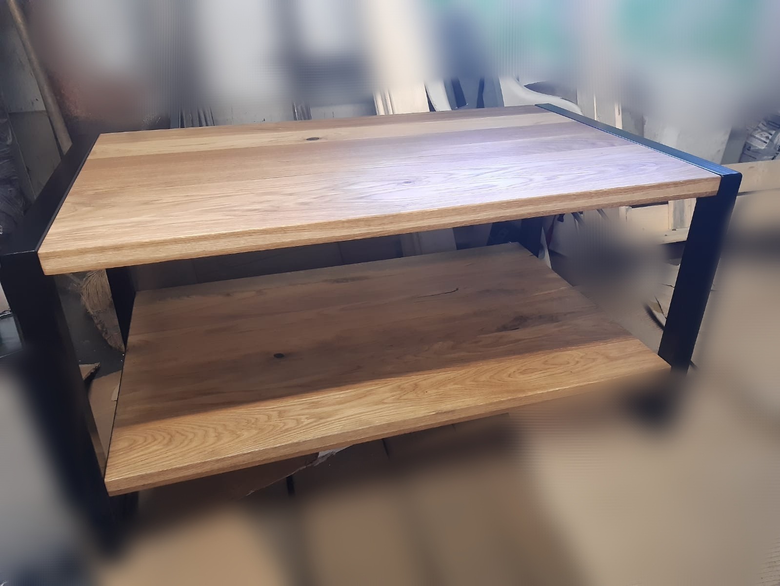 Stolik kawowy z dwoma blatami z jasnego drewna, osadzony na czarnych, metalowych nogach o prostokątnym przekroju. Widoczne usłojenie drewna i matowe wykończenie.