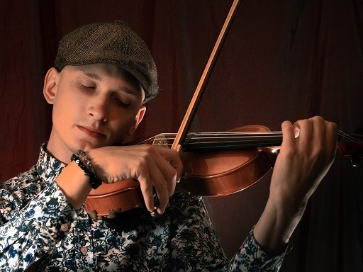 Młody mężczyzna w kaszkiecie, z zamkniętymi oczami, gra na skrzypcach, ubrany w koszulę w kwiaty, na ciemnym tle.