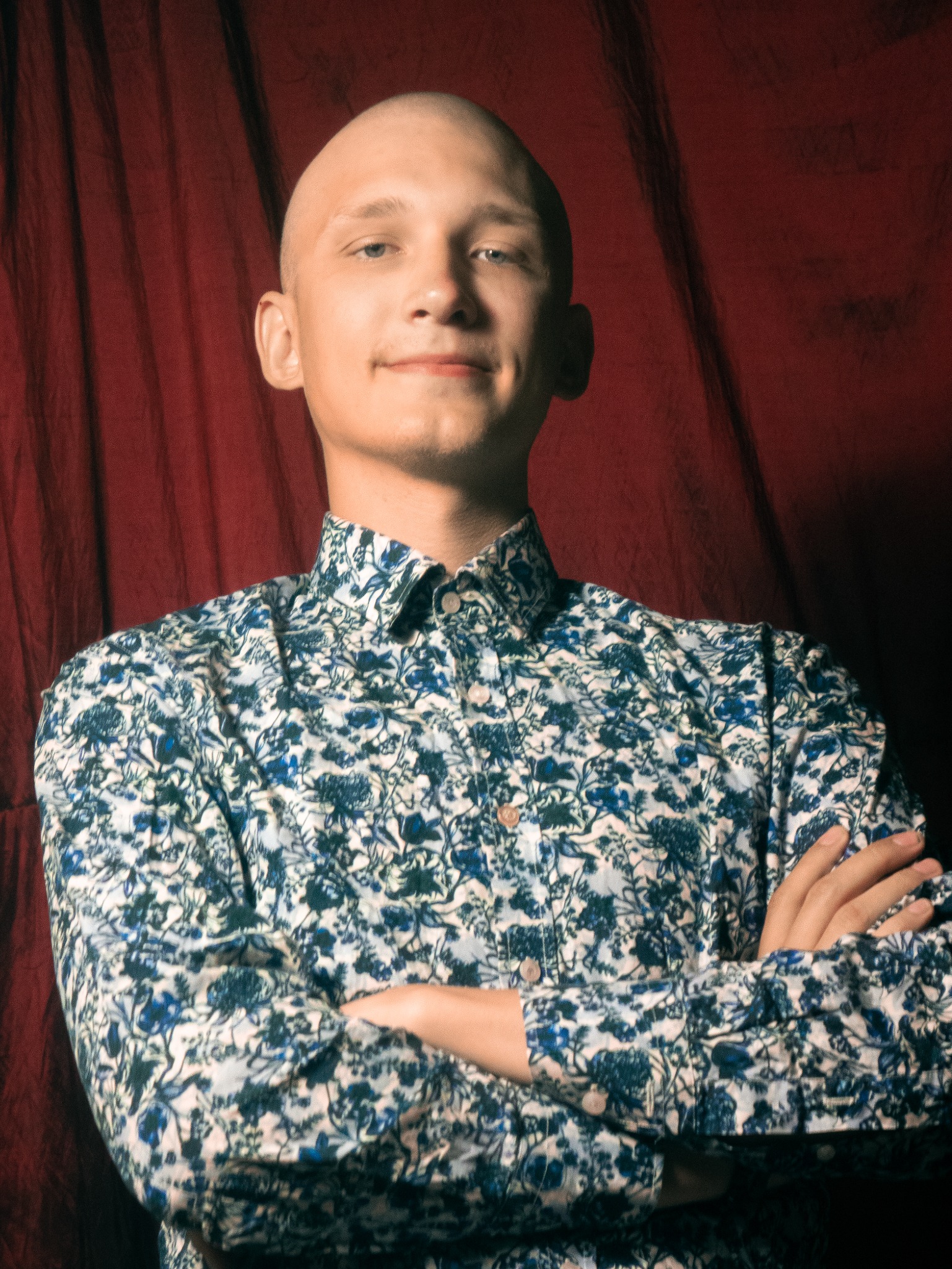 Portret młodego mężczyzny z ogoloną głową, ubranego w koszulę w niebieskie kwiaty, na tle czerwonej zasłony.