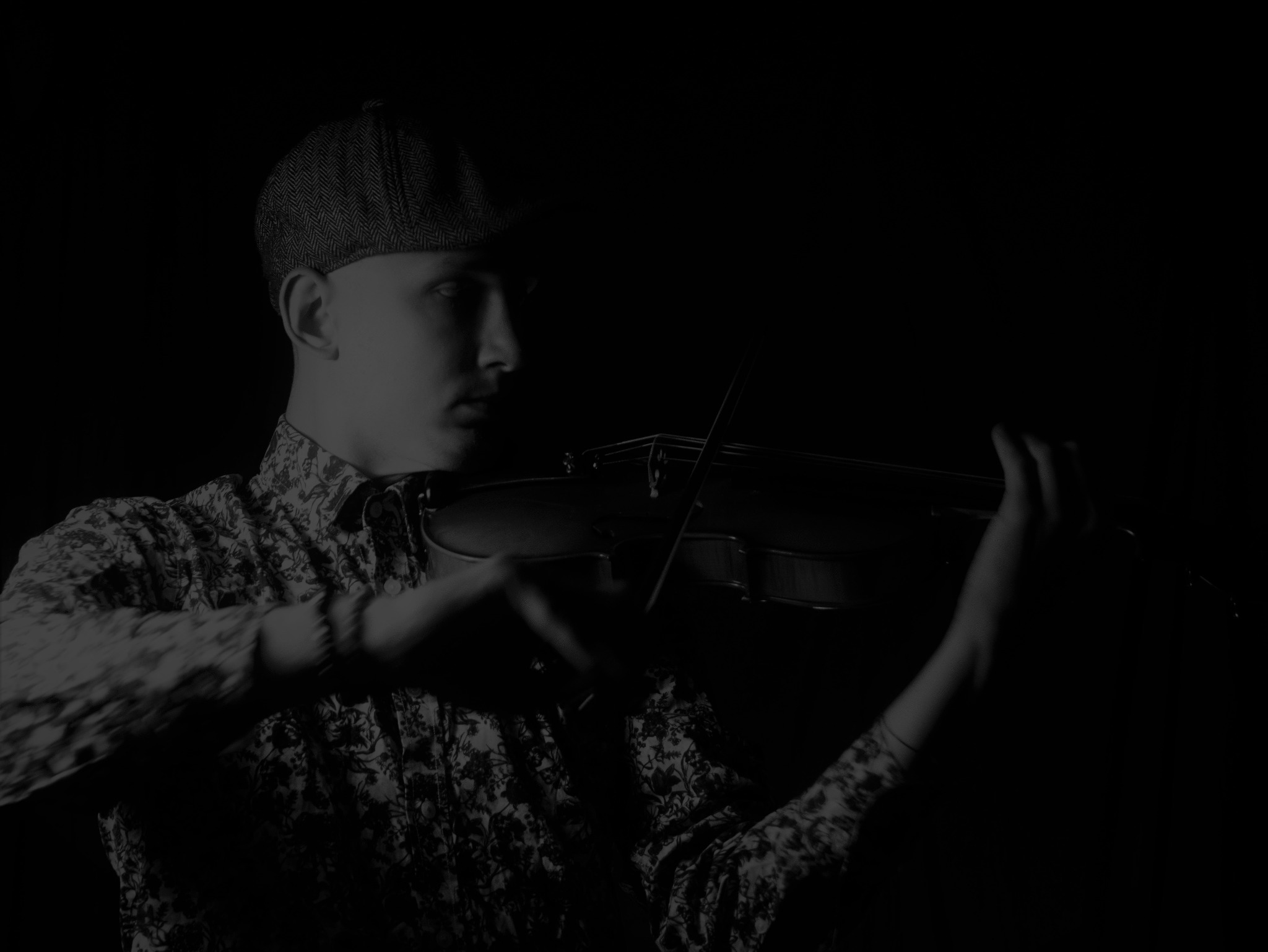 Młody mężczyzna w kaszkiecie i wzorzystej koszuli gra na skrzypcach w ciemnym otoczeniu, skupiony wzrok skierowany w lewo.