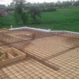 Zbrojenie płyty fundamentowej z drewnianym szalunkiem i izolacją termiczną, widok z góry na tle zielonego wzgórza z zabudowaniami.